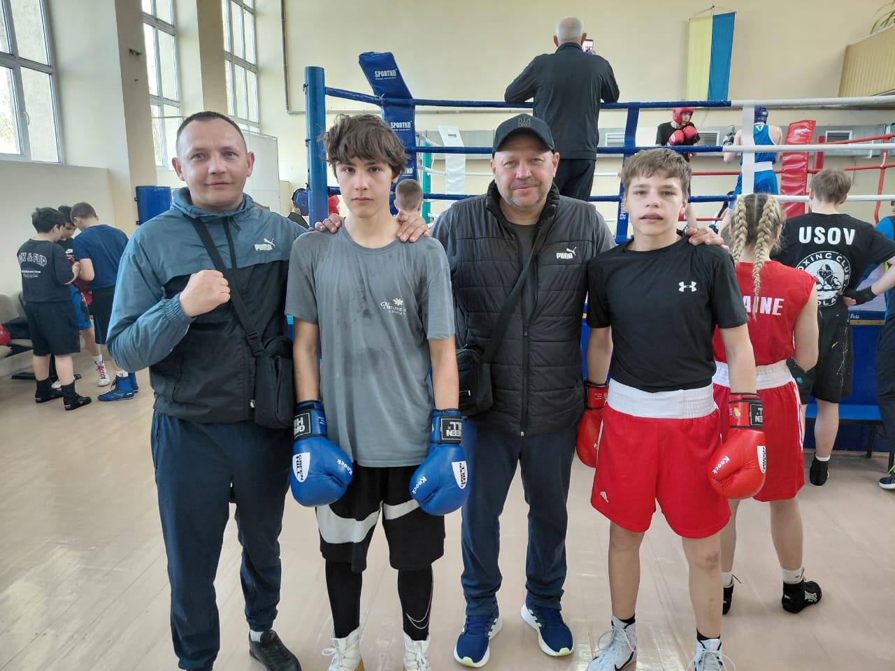 Четверо людей — двоє дорослих чоловіків та двоє юних спортсменів у боксерських рукавицях — позують у залі. На задньому плані видно боксерський ринг із канатами, спортсменів під час тренування та прапор України на стіні.