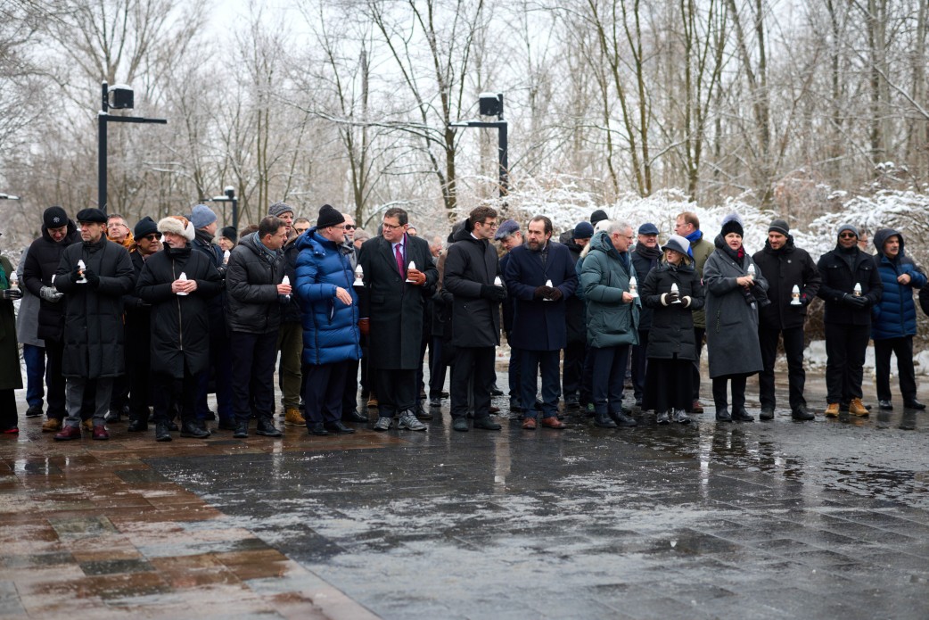 Велика група людей у зимовому одязі стоїть на засніженій площі під час меморіального заходу. Учасники тримають у руках запалені білі лампадки, вшановуючи пам'ять загиблих.