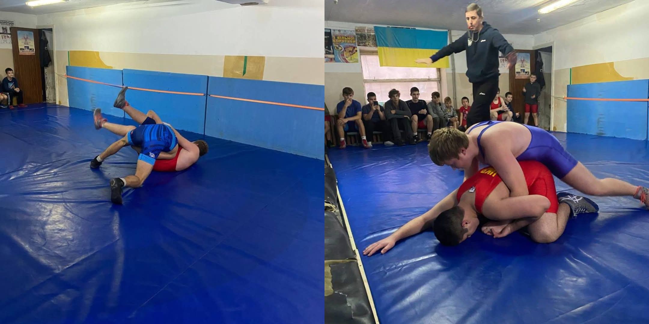 Це горизонтальний колаж, що складається з двох фотографій, які відображають моменти боротьби на тренуванні чи змаганнях у спортивному залі. Обидві сцени відбуваються на синьому борцівському килимі. Стіни залу світлі, нижня частина закрита синіми матами. Ліва частина: Двоє борців під час поєдинку. Один спортсмен у червоному трико лежить на спині, а інший у синьому трико, використовуючи прийом, перекидає його через себе. Права частина: Двоє борців у процесі поєдинку. Спортсмен у синьому трико знаходиться зверху, притискаючи спортсмена у червоному трико до килима. На задньому плані стоїть тренер (або суддя) у чорній спортивній кофті, який спостерігає за боротьбою, витягнувши руку вбік. Далі, вздовж стіни, сидять інші спортсмени та глядачі. На стіні видно жовто-блакитний прапор (прапор України) та плакати. Короткий опис: Колаж із двох знімків, що демонструють активні моменти боротьби між спортсменами у синьому та червоному трико на синьому борцівському килимі в спортивному залі під наглядом тренера/судді.