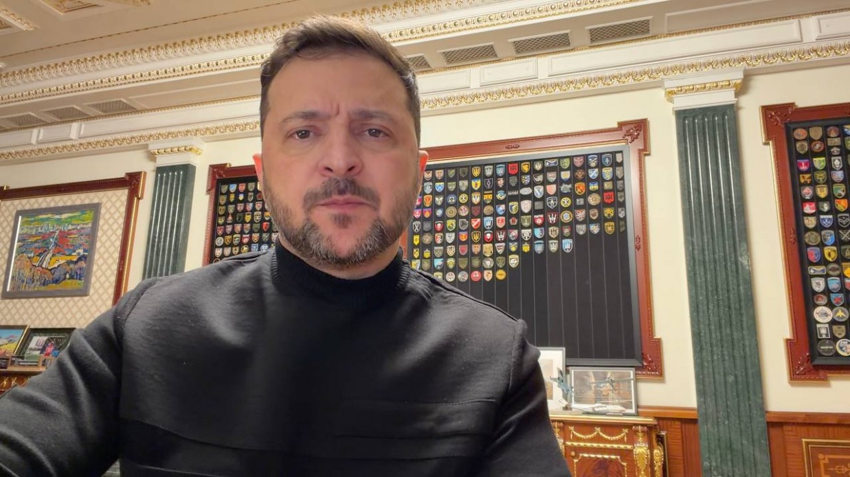 Чоловік у чорному светрі звертається до глядачів на фоні класичного офісного інтер’єру. Позаду нього на стіні розміщені стенди з численними військовими шевронами та підрозділовими емблемами, а також картина у рамі та декоративні колони.