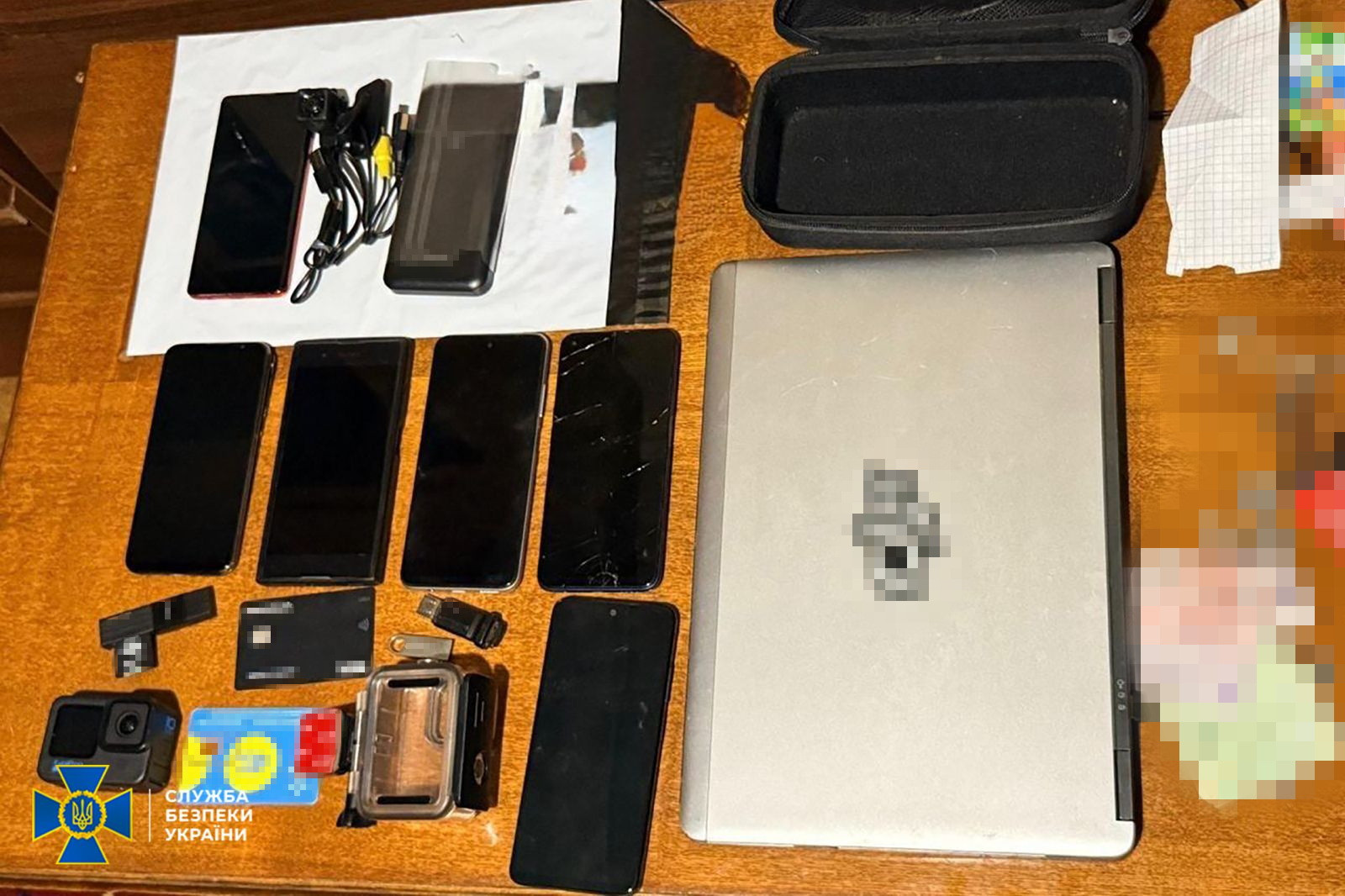 На дерев’яному столі розкладено вилучені речові докази: ноутбук, шість смартфонів, екшн-камеру, банківські картки, флеш-накопичувачі та зарядні пристрої. У нижньому лівому куті розміщено логотип Служби безпеки України.