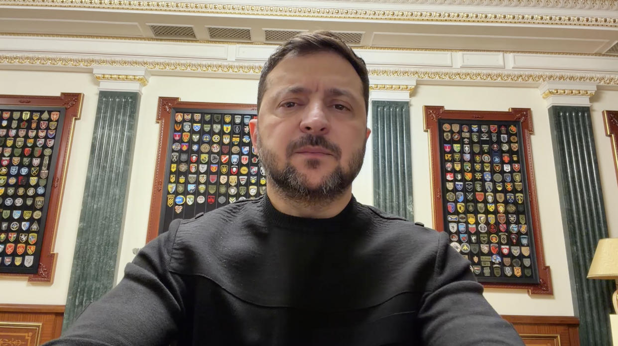 Чоловік у чорному светрі звертається до глядачів на фоні офіційного інтер'єру з колонами та декоративними рамками, заповненими численними військовими шевронами.