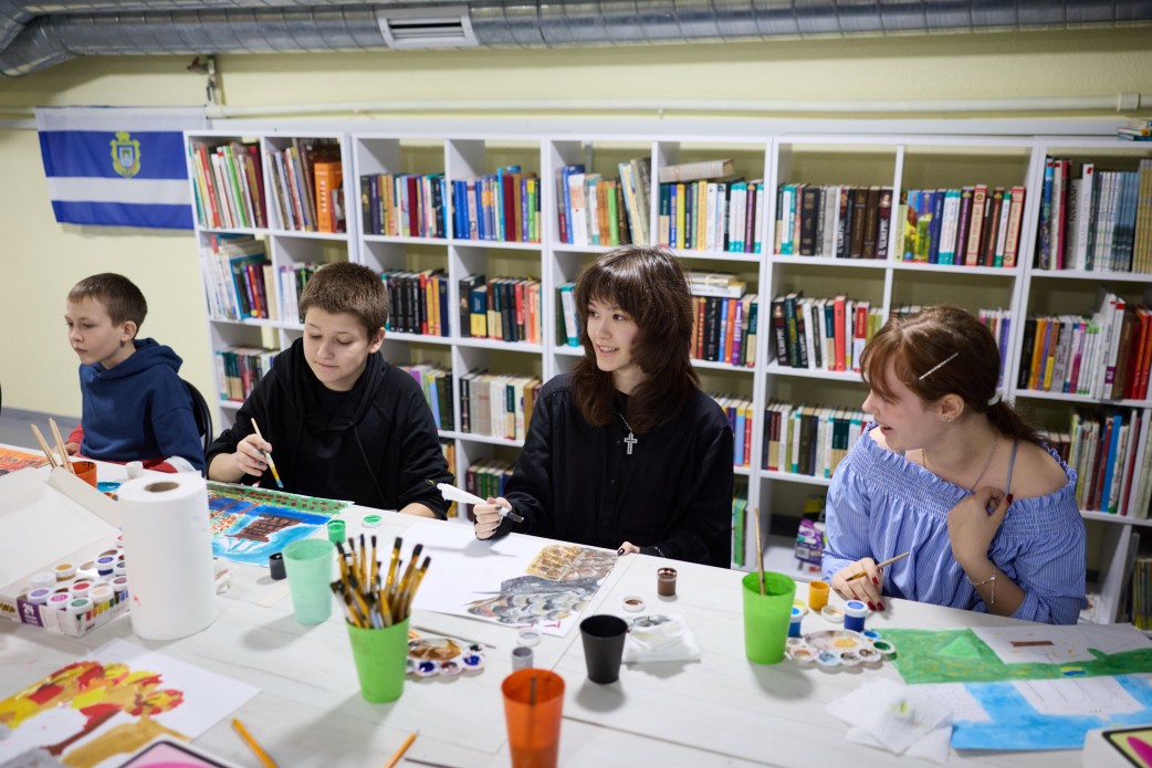 Четверо підлітків (двоє хлопців і дві дівчини) сидять за столом і займаються малюванням. На столі видно фарби, пензлі та їхні роботи. За ними розташований великий білий стелаж, заповнений книгами. На стіні ліворуч висить прапор.