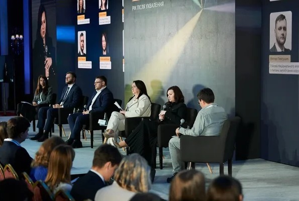 Панельна дискусія на конференції, де шість спікерів сидять у кріслах на сцені перед аудиторією на тлі темної стіни з портретами та написом «Рік після ухвалення».