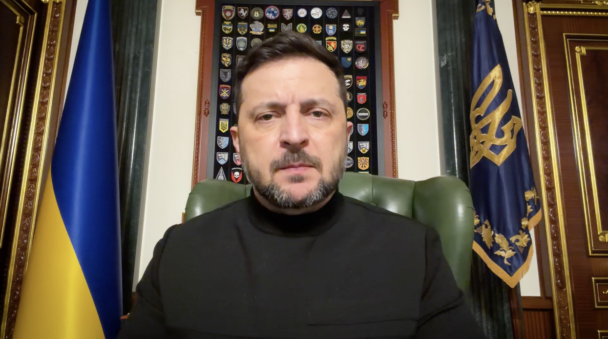 Чоловік у чорному одязі виступає на фоні державного прапора України та прапора з гербом. Позаду нього на стіні розміщено стенд із колекцією військових шевронів. Він сидить у шкіряному кріслі в офіційному кабінеті з класичним інтер’єром.