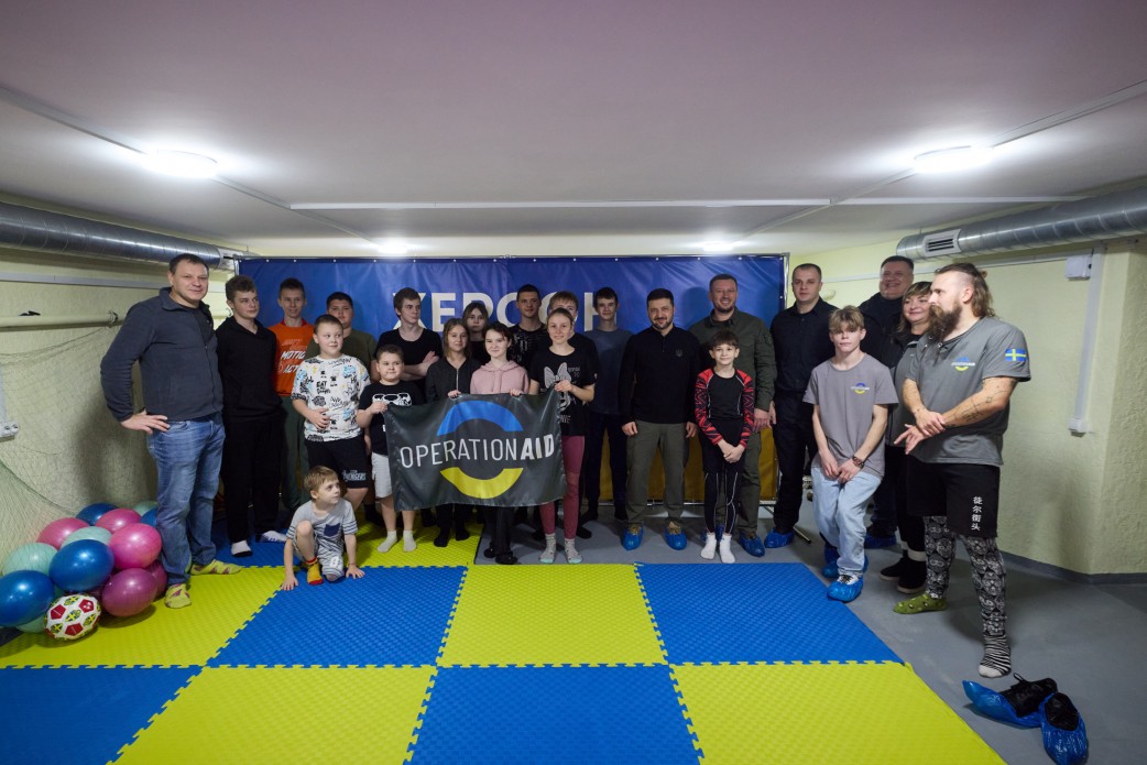 Групове фото Президента України Володимира Зеленського та його супроводу з групою підлітків та дітей. Учасники стоять у спортивному залі, тримаючи банер з написом: OPERATION AID. На підлозі лежать синьо-жовті мати та повітряні кульки.