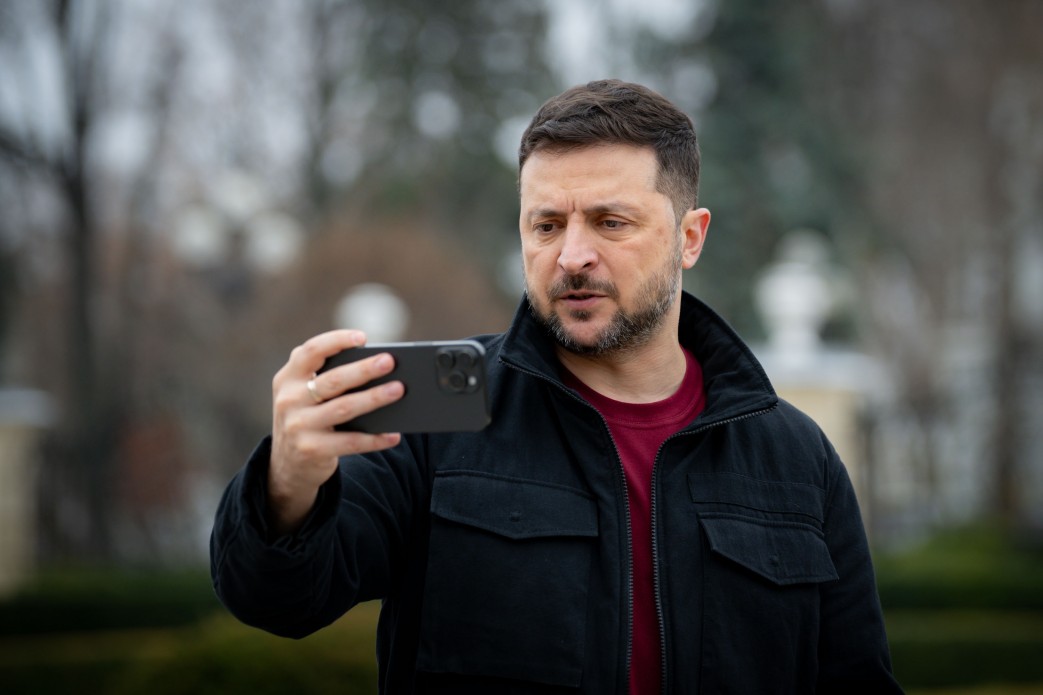 Крупний план Президента України Володимира Зеленського на вулиці, одягненого в чорну куртку та червону футболку, який тримає мобільний телефон горизонтально перед собою, ймовірно, для відеозапису чи дзвінка. Фон розмитий, видно дерева та елементи архітектури.