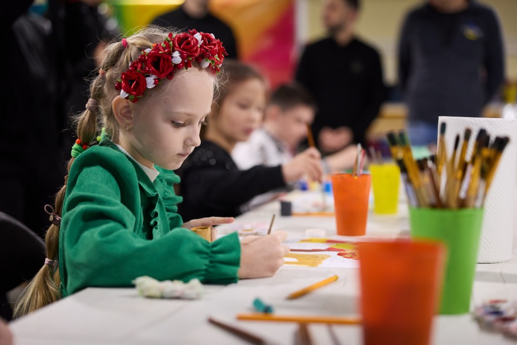 Крупний план дівчинки у вінку з червоних і білих квітів та зеленій кофтинці, яка сидить за столом і малює пензлем. На столі видно склянки з пензлями та фарби. На задньому плані в фокусі видно інших дітей, які також малюють, та силуети дорослих.