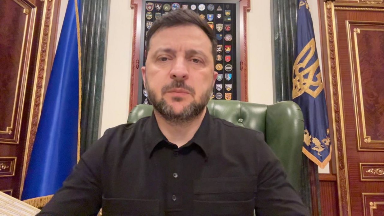 Президент України Володимир Зеленський у чорній сорочці сидить за столом у своєму кабінеті на тлі синьо-жовтих прапорів і вішака з емблемами.