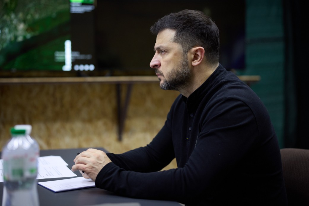 Крупний план Президента України Володимира Зеленського в чорній кофті, який сидить за столом у профіль, уважно слухаючи або розмірковуючи під час наради. Перед ним лежать документи, а на столі видно пляшку води.