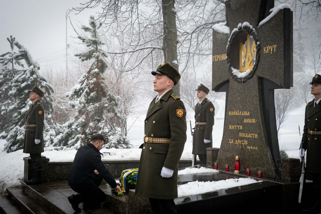Церемонія вшанування пам'яті біля меморіалу «Героям Крут». Чоловік у темному одязі покладає квіти до підніжжя пам'ятника у формі великого кам'яного хреста з тризубом. Поряд стоять почесна варта у парадній військовій формі. Навколо засніжений парк та дерева.
