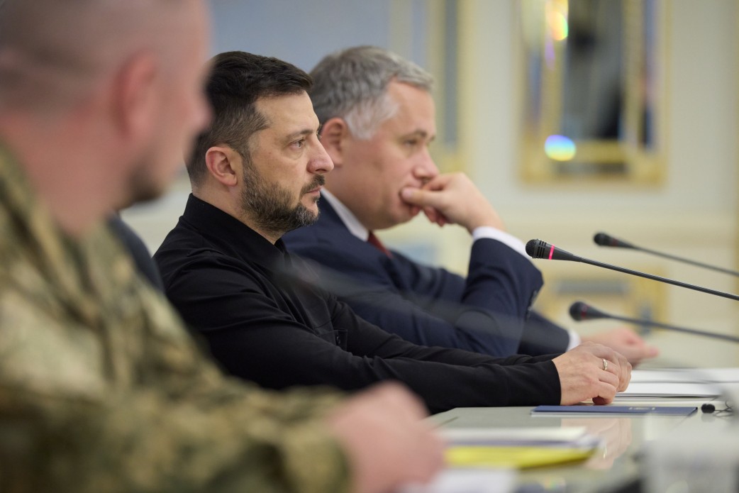 Чоловік у чорній сорочці сидить за столом під час офіційної зустрічі, зосереджено дивлячись перед собою. На фоні видно іншого учасника зустрічі в діловому костюмі та частину плеча військовослужбовця в камуфляжі на передньому плані.