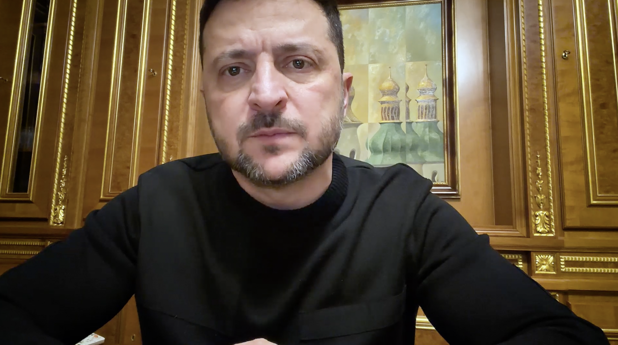 Чоловік із короткою борідкою, одягнений у чорний світшот, дивиться прямо в камеру. Він зафіксований крупним планом на фоні дерев'яних настінних панелей із золотим оздобленням та картини із зображенням церковних куполів.