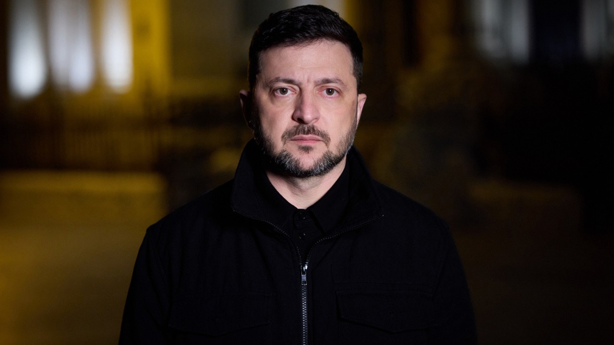 Портретний знімок Президента України Володимира Зеленського, який стоїть на вулиці вночі або в темний час доби. Він дивиться прямо в камеру, одягнений у чорну куртку на блискавці та сорочку-поло. На задньому плані — розмиті вогні.