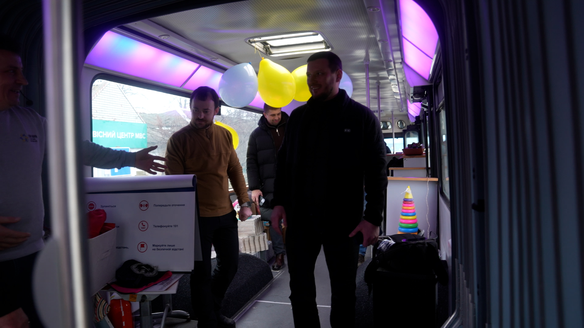 Двоє чоловіків перебувають усередині облаштованого автобуса з неоновим підсвічуванням та повітряними кульками. Один із них розглядає інформаційний стенд, інший стоїть поруч; на задньому плані видно дитячу пірамідку та інші речі для дозвілля.