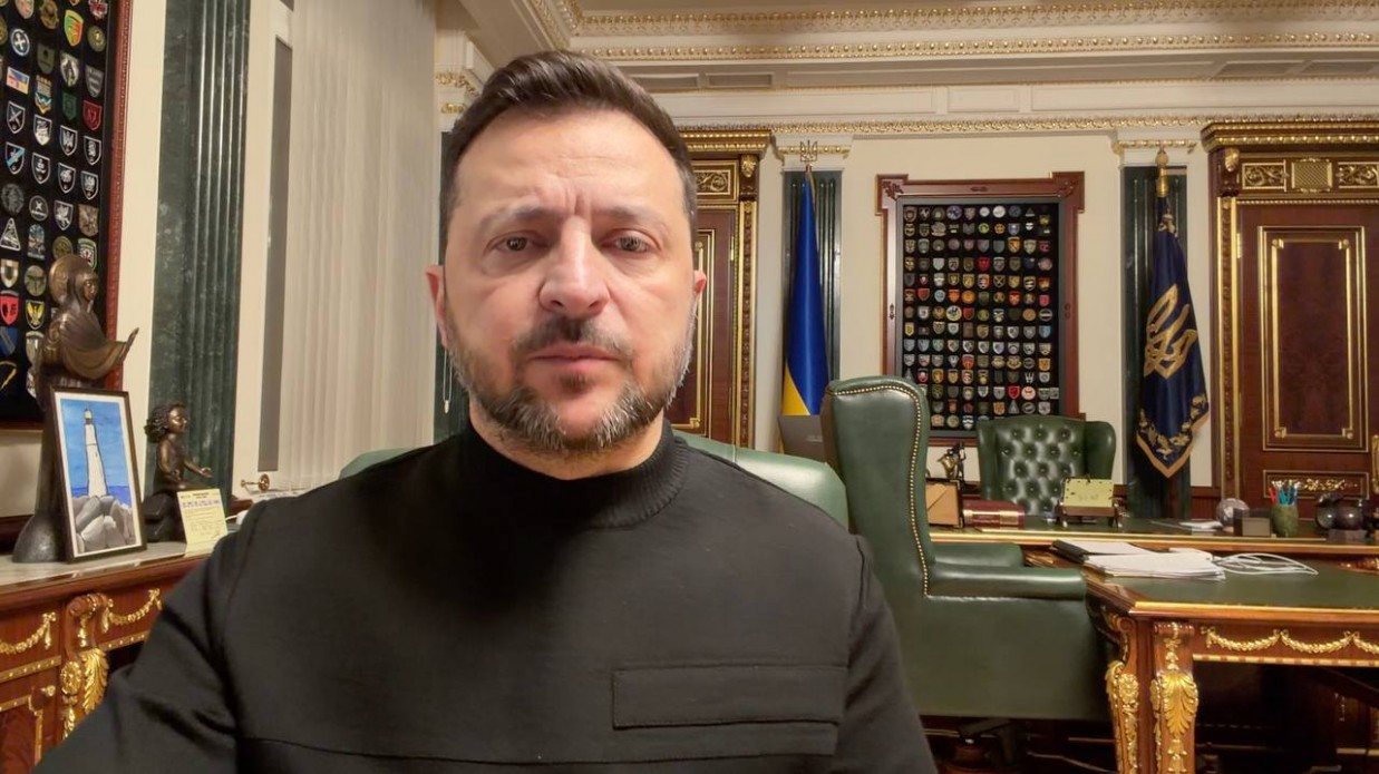 Чоловік у чорному одязі на фоні робочого кабінету з державними символами України. За ним видно робочий стіл, прапори та стіни, прикрашені численними шевронами військових підрозділів.