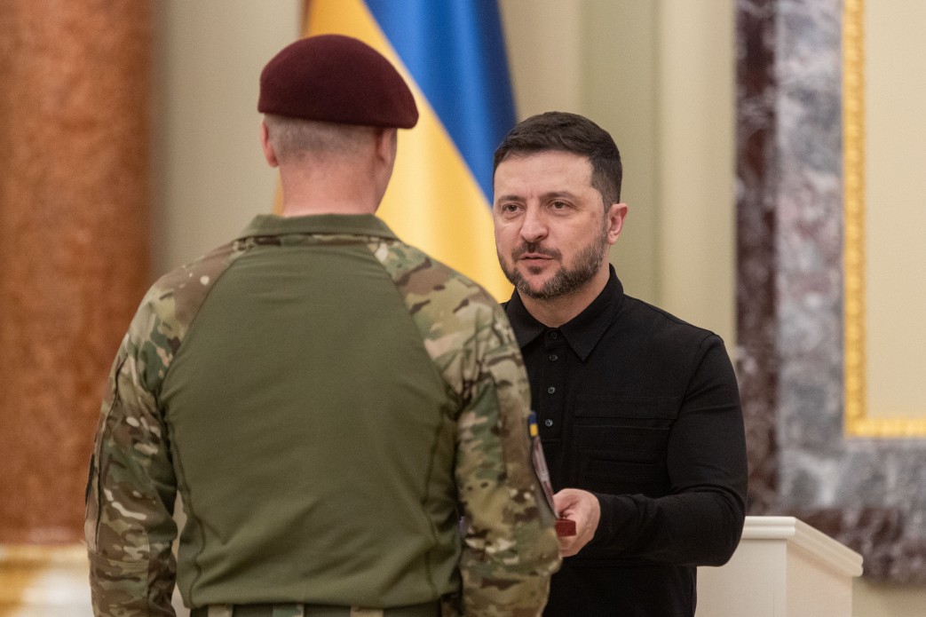 Президент України Володимир Зеленський у чорній сорочці вручає нагороду військовослужбовцю у камуфляжній формі та бордовому береті. На задньому плані видно прапор України.