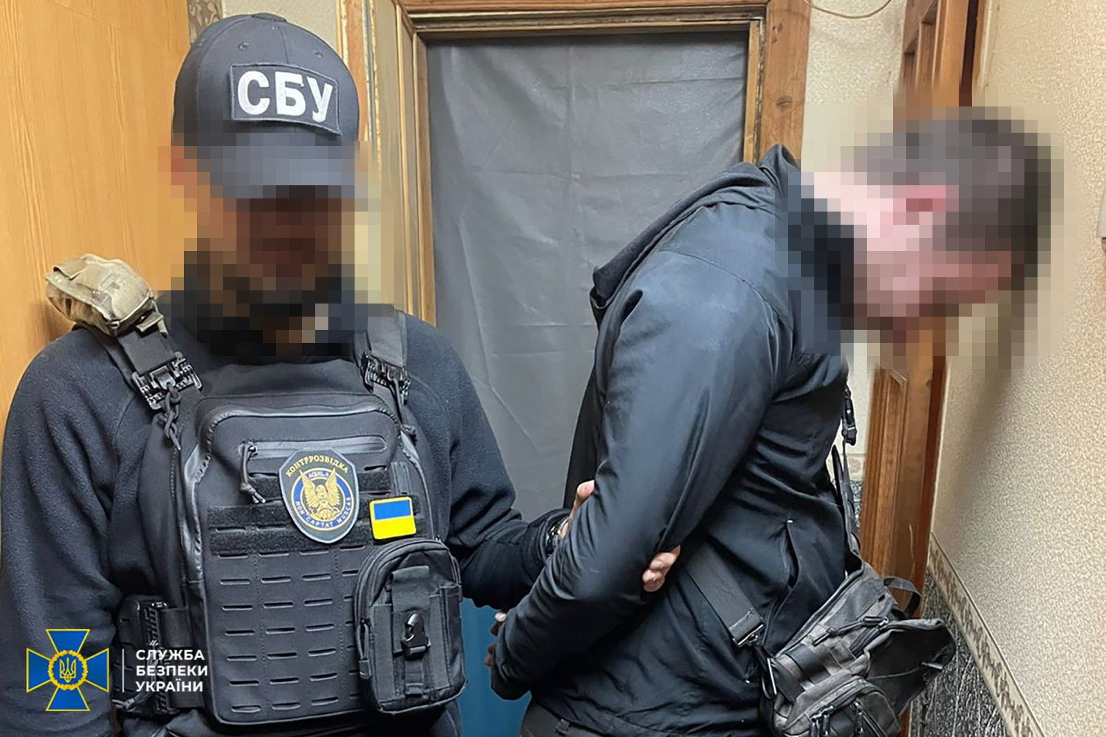 Затримання підозрюваного правоохоронцями. Ліворуч зображений співробітник контррозвідки СБУ у чорній тактичній кепці з написом «СБУ» та розвантажувальному жилеті з емблемою підрозділу і маленьким прапором України. Його обличчя закрите розмиттям. Він тримає під руку затриманого чоловіка у чорній куртці з капюшоном та з тактичною сумкою через плече. Затриманий стоїть спиною до камери, нахиливши голову вперед до стіни; його обличчя розмите пікселями. У лівому нижньому кутку нанесено емблему СБУ.