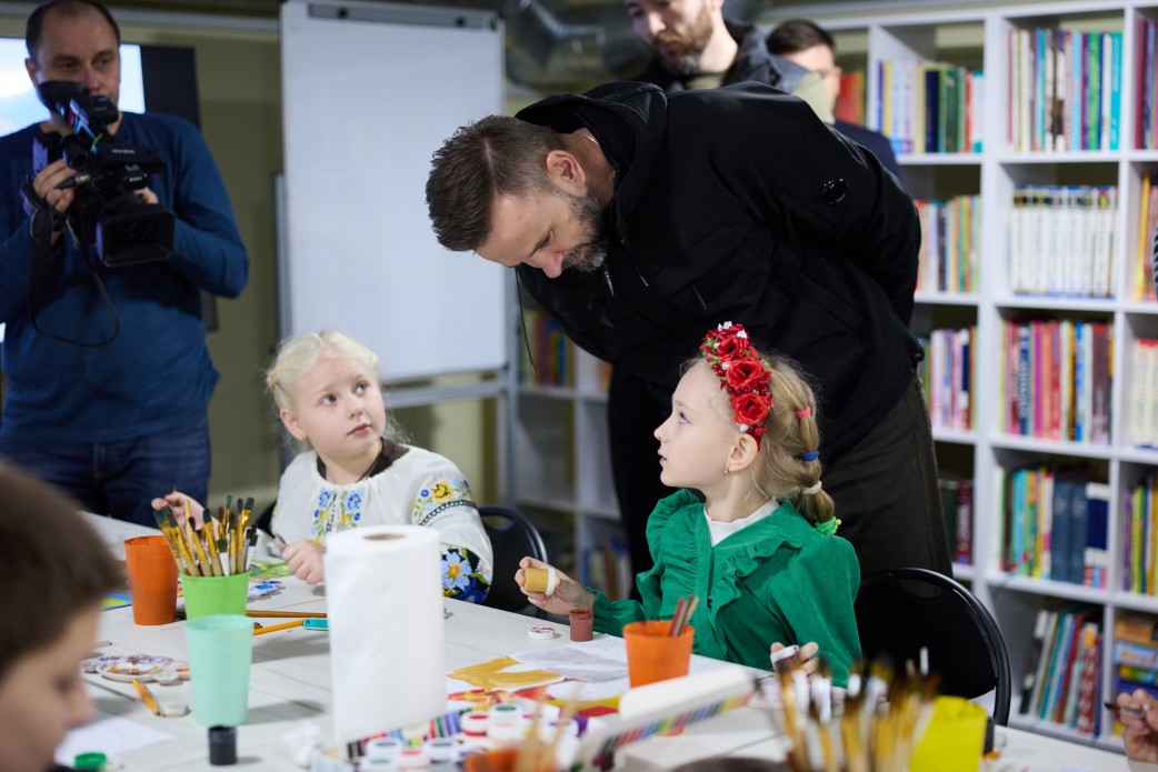 Чоловік у чорній куртці схиляється над столом, розмовляючи з двома дівчатками, які малюють. Одна дівчинка одягнена у вишиванку, інша — у зелену сукню і віночок з червоних троянд. На задньому плані видно книжковий стелаж та оператора з камерою.