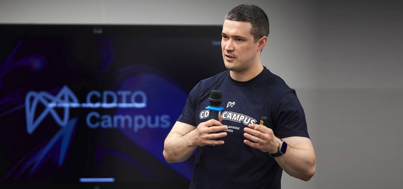 На зображенні чоловік проводить публічний виступ, тримаючи мікрофон у правій руці та пристрій для презентації в лівій. Він одягнений у темну футболку з логотипом «CDTO Campus». На задньому плані видно великий екран із таким самим логотипом і темно-фіолетовим фоном. Сцена освітлена м’яким світлом, що створює ділову та зосереджену атмосферу заходу, присвяченого підготовці цифрових лідерів.