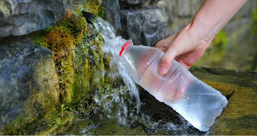 Крупний план, на якому видно, як рука людини набирає воду з кам'яного джерела (джерела) у прозору пластикову пляшку. Вода витікає з невеликої трубки або отвору в покритому мохом камені.