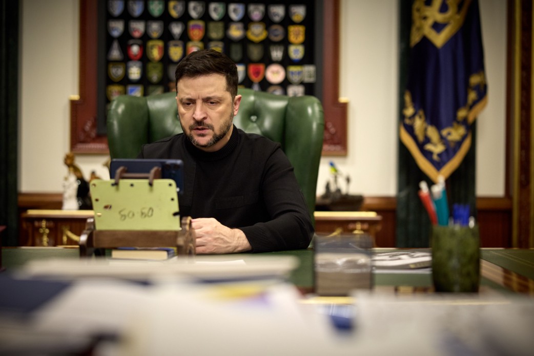 Чоловік у чорному одязі сидить за робочим столом у кабінеті та зосереджено дивиться на екран смартфона, встановленого на підставці. На фоні за його спиною розташоване зелене шкіряне крісло, державний прапор та стенд із великою колекцією військових шевронів.