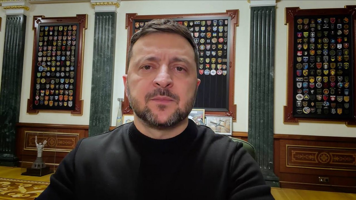 Чоловік у чорному одязі на фоні стіни з багатьма військовими шевронами у рамках та високими колонами. Він дивиться прямо в камеру під час офіційного відеозвернення.