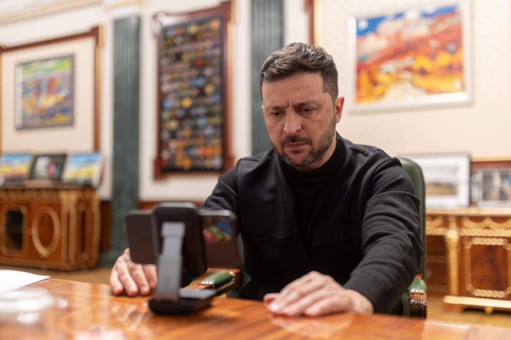 Володимир Зеленський сидить за полірованим столом у робочому кабінеті, дивлячись на смартфон, закріплений на підставці. На задньому плані видно картини.