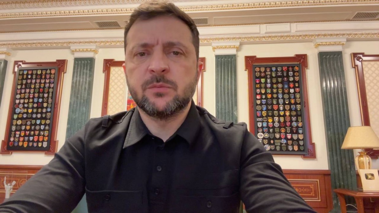 Президент України Володимир Зеленський у чорній сорочці сидить перед камерою у своєму робочому кабінеті під час нічного відеозвернення. На задньому плані видно багатий інтер'єр з різьбленими дерев'яними елементами, колонами та двома великими вертикальними вітринами, заповненими численними військовими шевронами та нашивками.