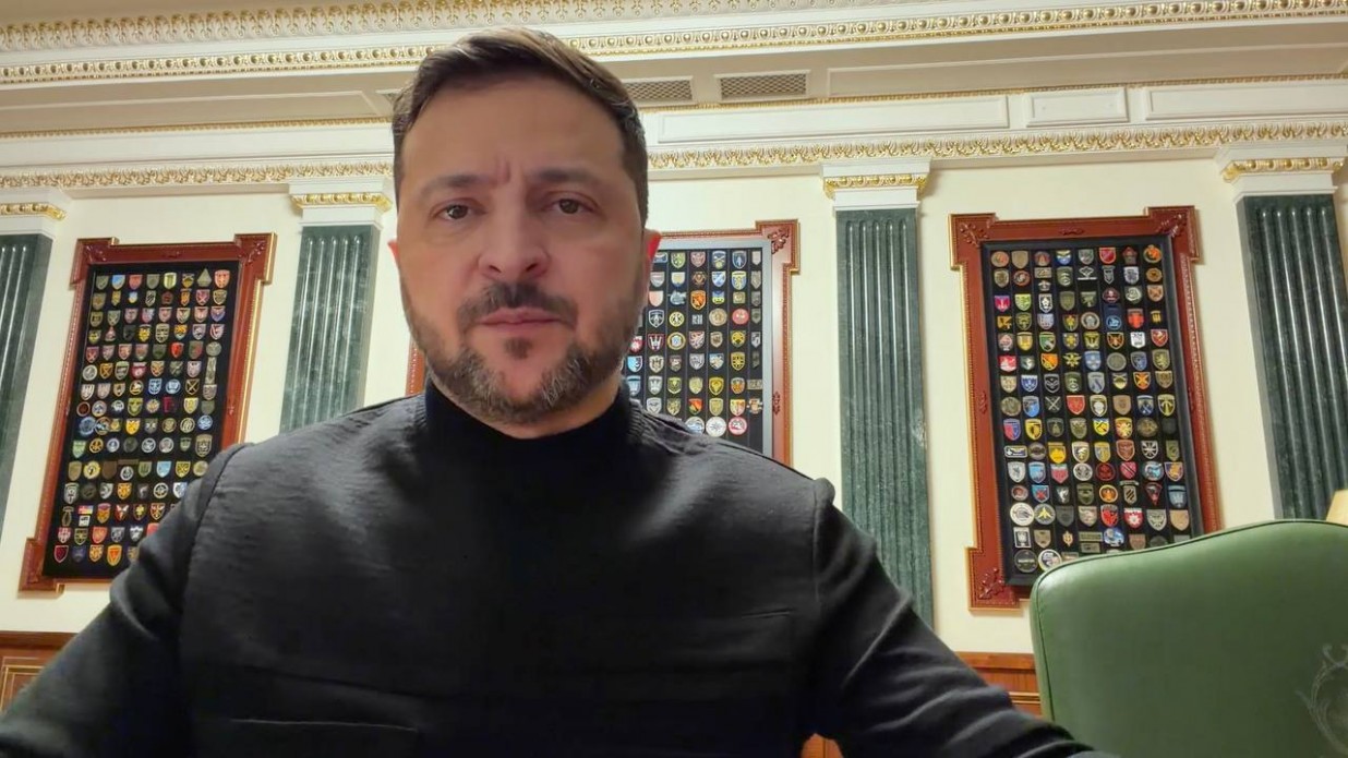 Чоловік із короткою бородою та в чорному гольфі стоїть на фоні офісного приміщення з колонами. Позаду нього на стіні розміщені три великі рамки з численними військовими шевронами та емблемами підрозділів.