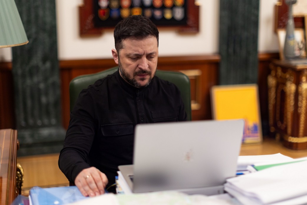 Чоловік у чорній сорочці сидить за робочим столом і зосереджено дивиться в екран відкритого ноутбука. Навколо нього лежать документи та папки, а на фоні видно класичний інтер'єр кабінету з дерев'яними елементами та прапорами.