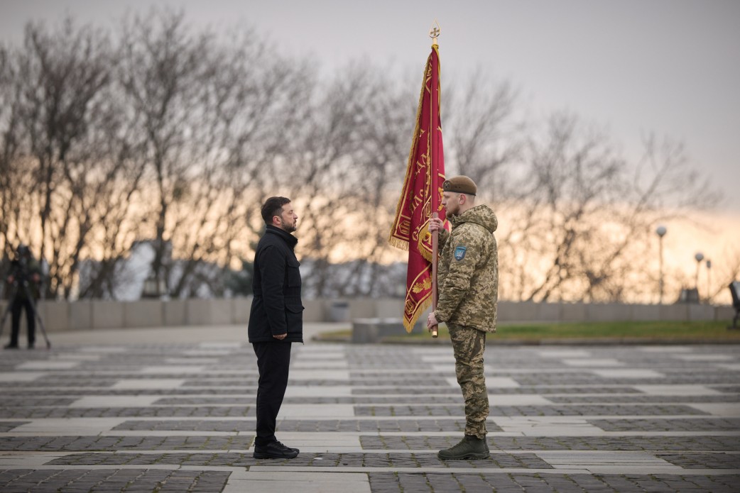 На фото зображений Президент України Володимир Зеленський у чорній куртці, який вручає нагороду військовослужбовцю в камуфляжній формі та оливковому береті. Дія відбувається на великій площі на тлі дерев і похмурого неба.