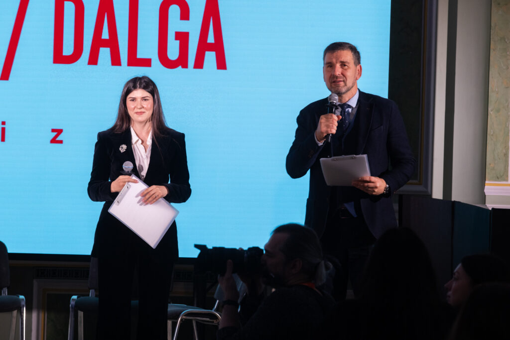 На сцені біля великого екрана з написом «DALĞA prize» стоять двоє ведучих — жінка та чоловік. Вони тримають мікрофони та папки з документами, звертаючись до аудиторії.