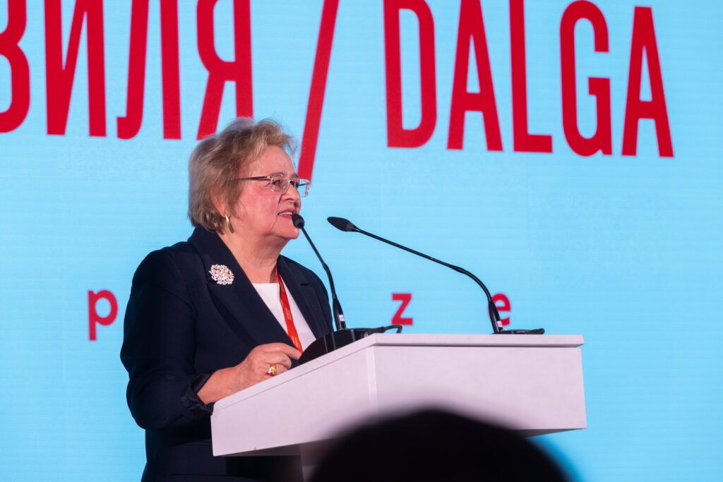 Жінка старшого віку в окулярах та темному жакеті з брошкою виступає за білою трибуною з мікрофонами. На задньому плані — світлий екран із червоними написами та частиною назви премії «DALĞA».