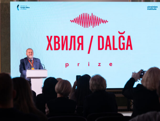 Чоловік виступає за трибуною на фоні великого екрана з написом «ХВИЛЯ / DALĞA prize». На передньому плані видно силуети глядачів у залі, деякі з яких знімають подію на телефони.
