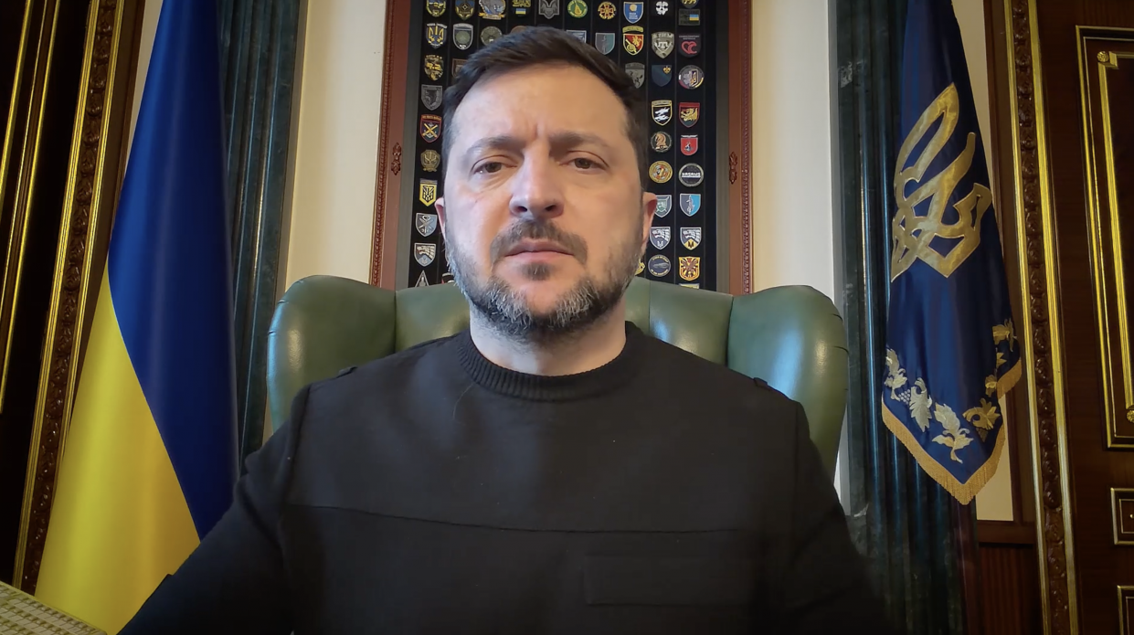 Чоловік у темному одязі виступає зі зверненням, сидячи в кріслі на офіційному фоні. Позаду нього розташовані Державний Прапор України та штандарт Президента України, а на стіні по центру розміщена дошка з шевронами різних військових підрозділів.