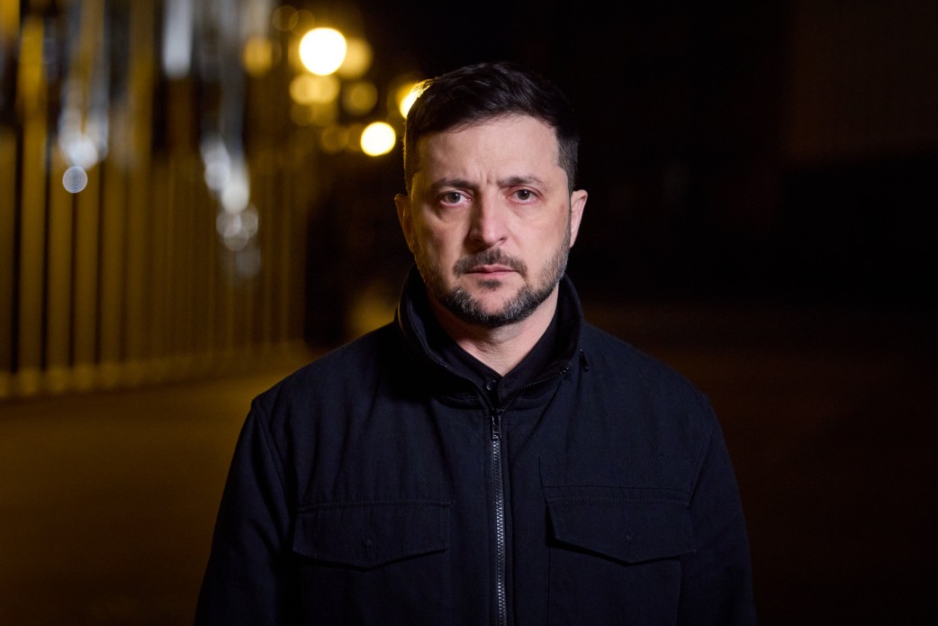 Крупний план Президента України Володимира Зеленського, який стоїть на вулиці вночі. Він одягнений у чорну куртку, має бороду і серйозний вираз обличчя. На задньому плані, що знаходиться поза фокусом, видно розмиті жовті вуличні ліхтарі.