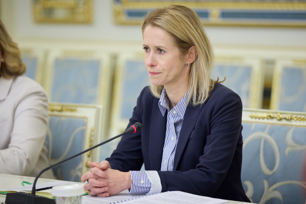 Жінка у темно-синьому жакеті та смугастій сорочці сидить за столом під час офіційної зустрічі. Вона зосереджено слухає, зчепивши пальці рук перед собою. На фоні видно частину класичного інтер’єру зали з декоративною позолотою та блакитними кріслами.