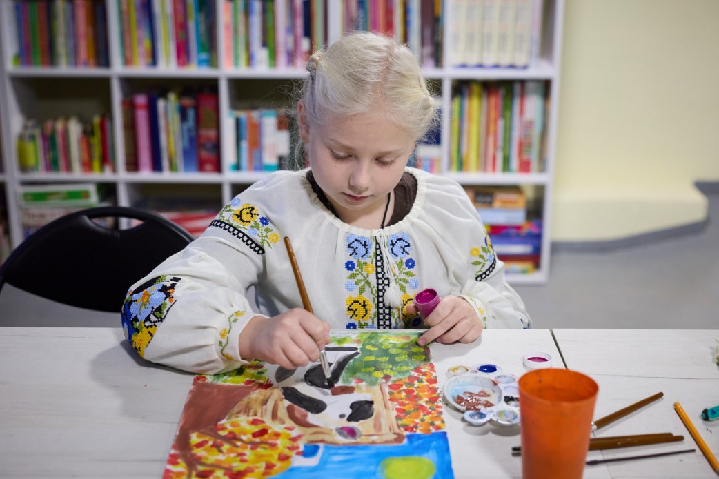 Дівчинка зі світлим волоссям, одягнена у вишиванку, сидить за столом і зосереджено малює пензлем аквареллю або гуашшю. На столі лежать фарби та пензлі. На задньому плані видно білий стелаж із кольоровими книгами.