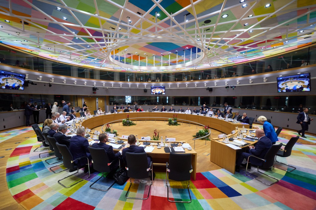 Ось короткий альтернативний текст для останнього зображення:  Панорамний вигляд великої зали засідань у будівлі Європейської Ради. Учасники офіційної зустрічі сидять за великим круглим столом у центрі зали, яка оздоблена яскравим різнокольоровим килимом та має складну геометричну стелю з підсвіткою.
