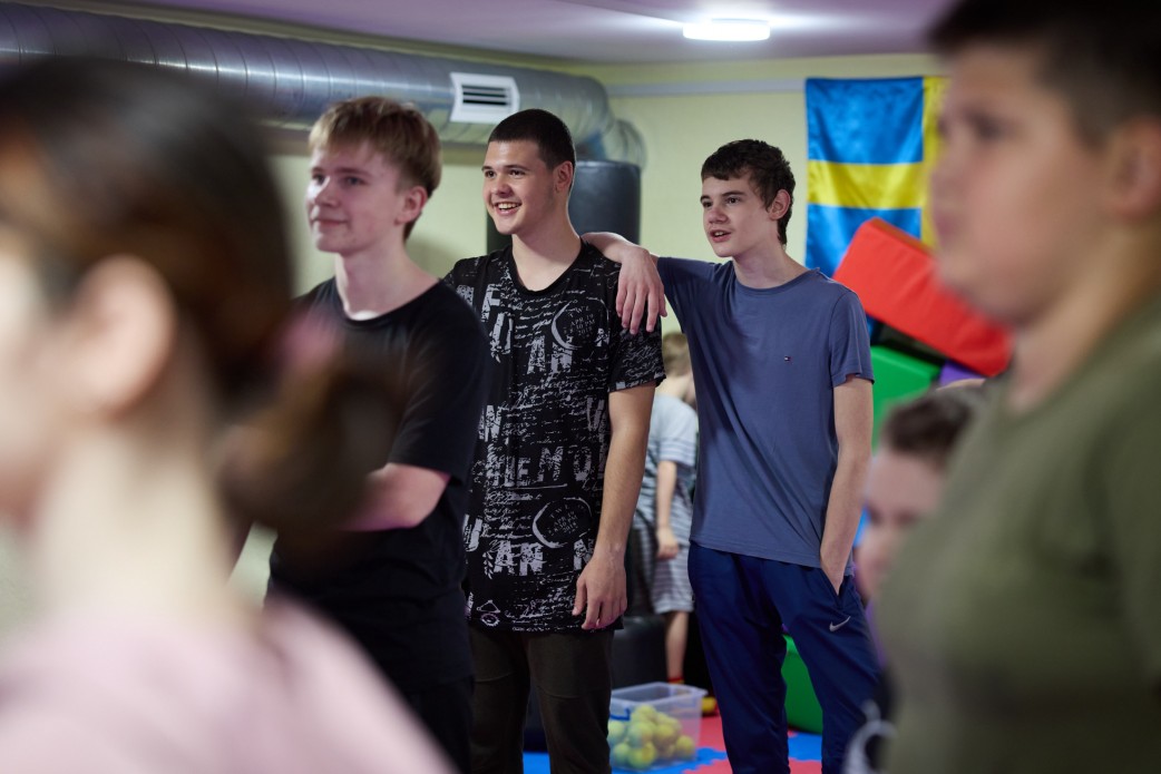 Троє усміхнених хлопців-підлітків стоять разом у приміщенні з м'якими ігровими модулями на задньому плані, де також видно синьо-жовтий прапор (можливо, прапор Херсонської області). Знімок зроблено через плече іншої людини на передньому плані.