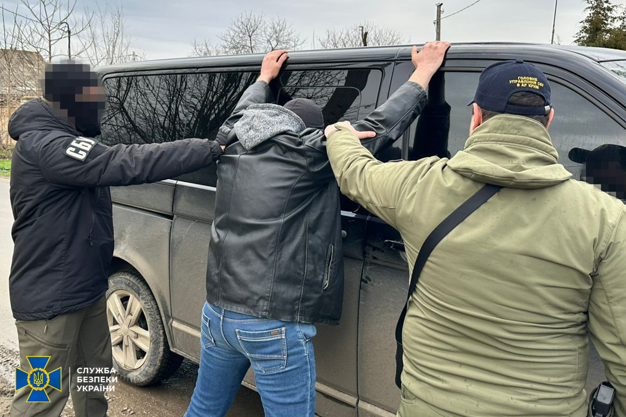 Двоє співробітників Служби безпеки України проводять затримання чоловіка. Затриманий стоїть спиною до камери, спершись руками на чорний мікроавтобус. Один із правоохоронців одягнений у куртку з написом «СБУ» та балаклаву, інший — у тактичний одяг із шевроном головного управління СБУ. Обличчя на фотографії розмиті для збереження анонімності. У нижньому куті розміщено логотип Служби безпеки України.