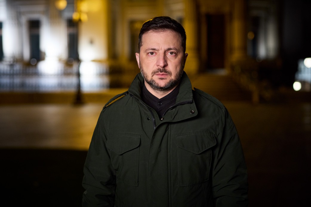 Портрет Президента України Володимира Зеленського, знятий вночі. Він стоїть, дивлячись прямо у камеру, одягнений у темно-зелену військову куртку та чорну сорочку. Фон розмитий (боке), видно лише неясні контури будівель і вуличних ліхтарів, що створює драматичне освітлення.