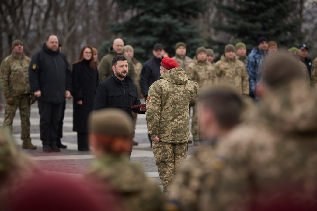 На фото зображено Президента України Володимира Зеленського, який стоїть в центрі кадру в чорній куртці, вручаючи нагороду військовослужбовцю в камуфляжній формі та червоному береті. Церемонія відбувається на вулиці, на задньому плані стоять інші військові та цивільні особи.