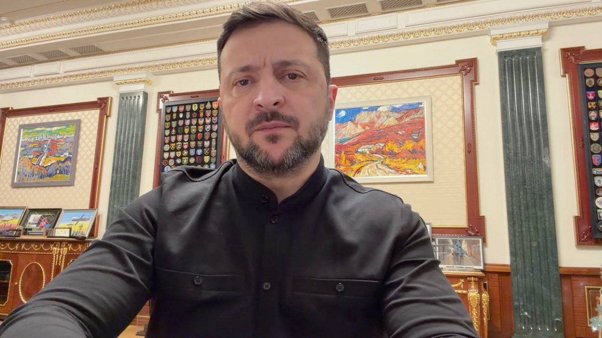 Чоловік із короткою бородою у чорній сорочці на фоні класичного офісного інтер’єру. На стінах за його спиною розміщені картини у декоративних рамах та стенди з емблемами (шевронами). Приміщення має високу стелю з ліпниною та колони.