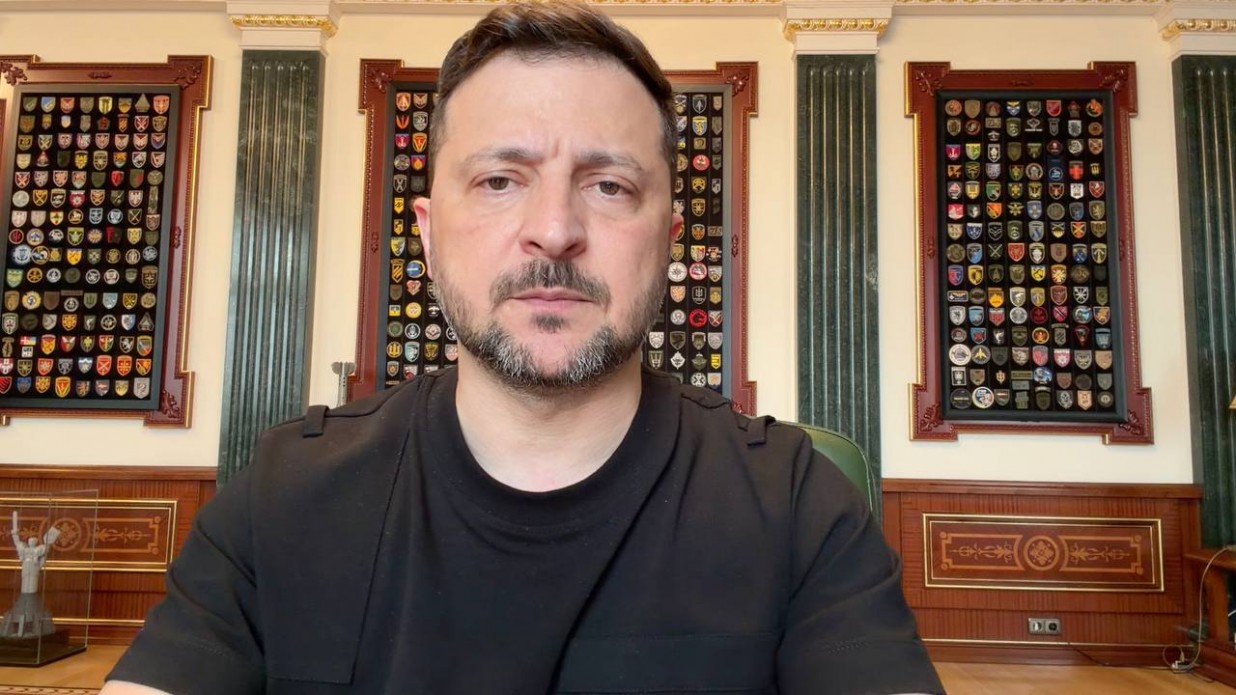 Володимир Зеленський великим планом під час звернення. Президент з короткою стрижкою, сивиною на скронях та акуратною борідкою дивиться в об'єктив із серйозним та втомленим виразом обличчя. На ньому чорна футболка з тактичними елементами (погонами) на плечах. Кадр зроблено в кабінеті: на стінах позаду розташовані три великі декоративні стенди в дерев'яних рамах, повністю заповнені різнокольоровими нарукавними знаками (шевронами) військових підрозділів України. Нижче видно дерев'яне оздоблення стін із класичним різьбленням.