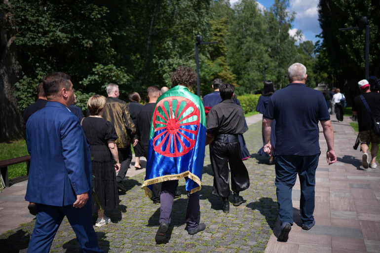 Група людей іде алеєю парку під час офіційного заходу. У центрі кадру людина несе на плечах синьо-зелений прапор із червоним колесом (прапор ромів). Учасники зняті зі спини, вони одягнені переважно в темний одяг.