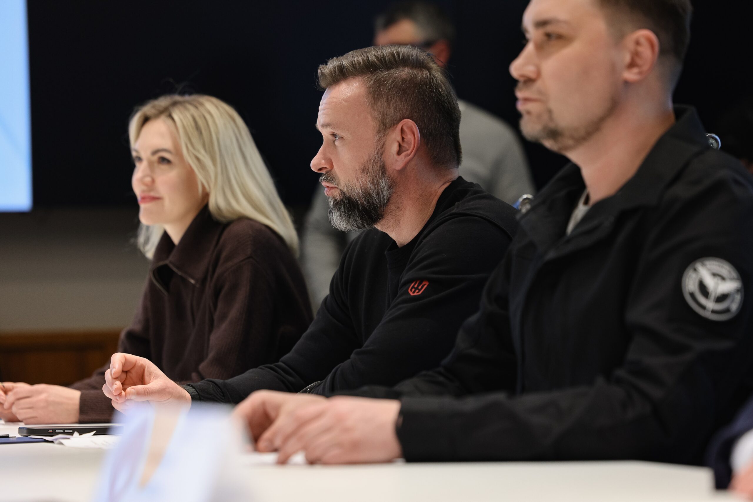 Троє людей у темному одязі сидять у ряд за довгим столом під час ділової зустрічі, дивлячись в одному напрямку. На передньому плані видно чоловіка у тактичній куртці з нашивкою, а поряд із ним — чоловіка з бородкою та жінку зі світлим волоссям.