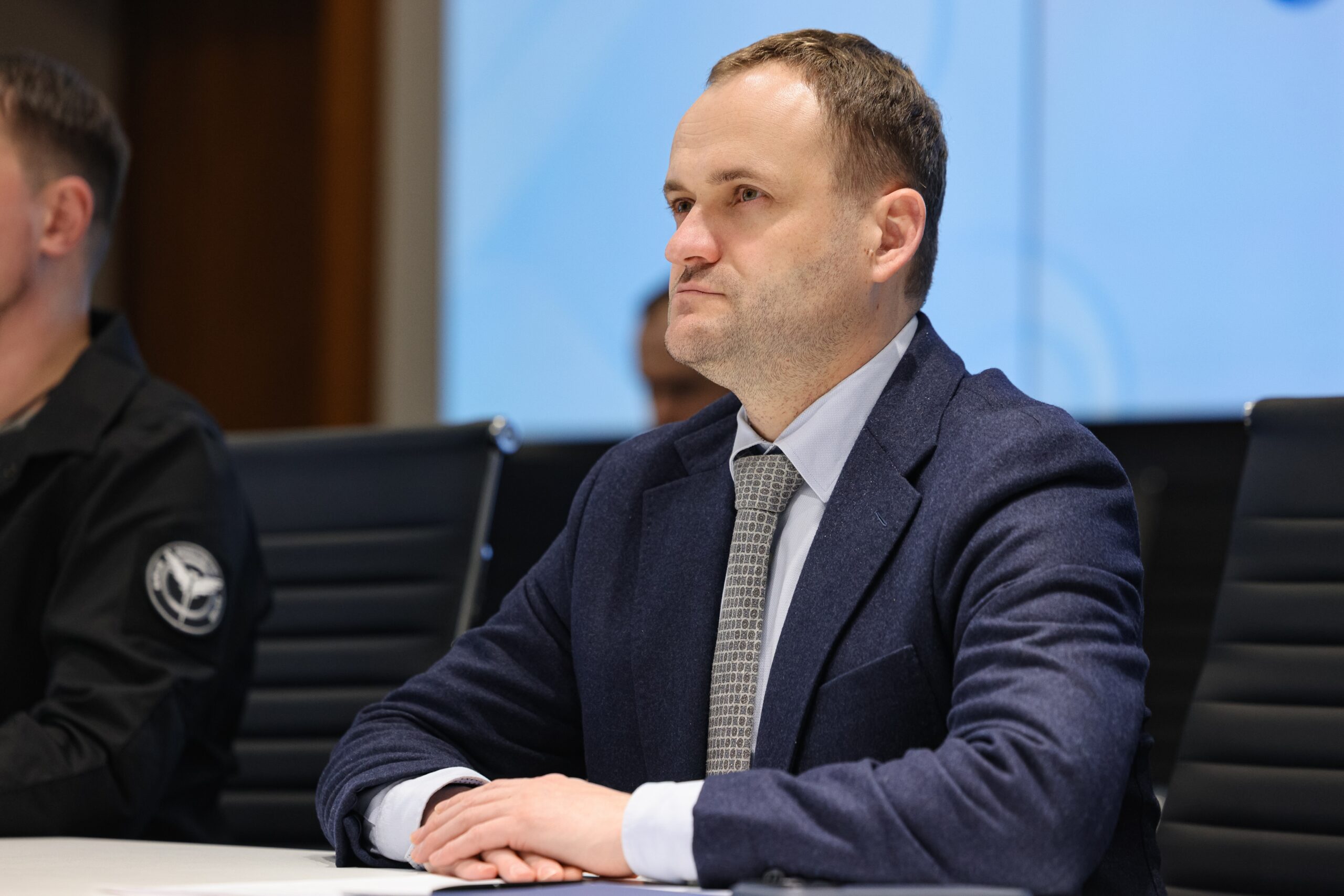 Чоловік у синьому піджаку, сорочці та краватці сидить за столом, склавши руки перед собою та дивлячись у бік. Він знаходиться в офіційному залі на фоні блакитного екрана, а поруч із ним видно частину плеча іншого учасника заходу в темному одязі з нашивкою.