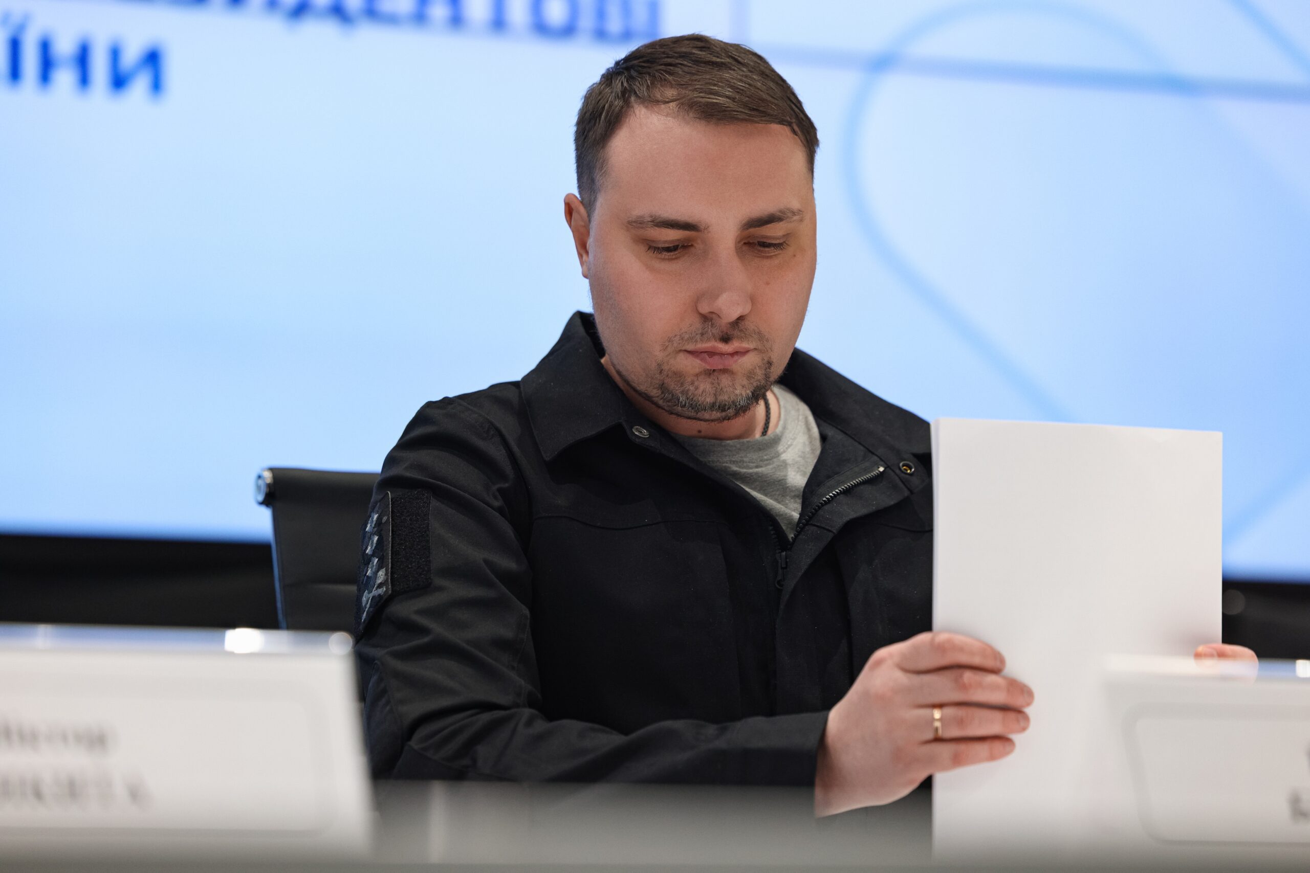 Чоловік у чорній тактичній куртці сидить за столом і зосереджено переглядає білий аркуш паперу. На фоні видно частину світлого екрана з офіційним написом, а перед чоловіком на столі розміщена ідентифікаційна табличка.
